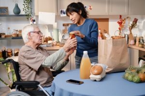 Volunteer delivering food to senior man using wheelchair, both sitting in the kitchen having a conversation