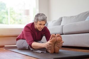 Beautiful senior woman doing stretching exercise while sitting on yoga mat at home. Mature woman exercising in sportswear by stretching forward to touch toes. Healthy active lady doing yoga and flexibility exercise.