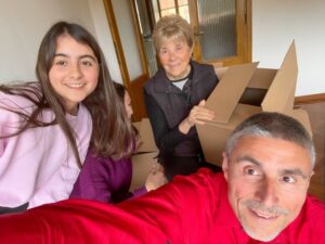 Moving to a new home concept. Young girl and her parents and grandmother seated near cardboard boxes along the  parquet floor taking selfies with smartphone
