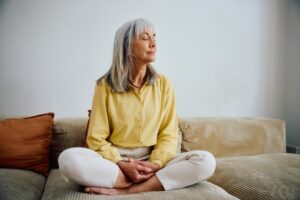 Full length of a relaxed senior woman sitting cross-legged on a beige sofa, meditating with her eyes closed and hands together inside a cozy home interior.